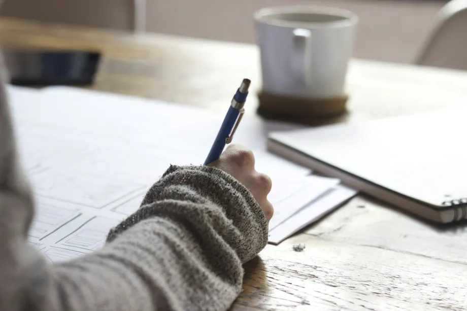 Person sketching page wireframes in a notebook on a wooden table beside a white mug